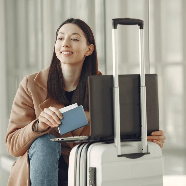 woman kneeling on the ground with her laptop open sitting on her suitcase and holding her passport