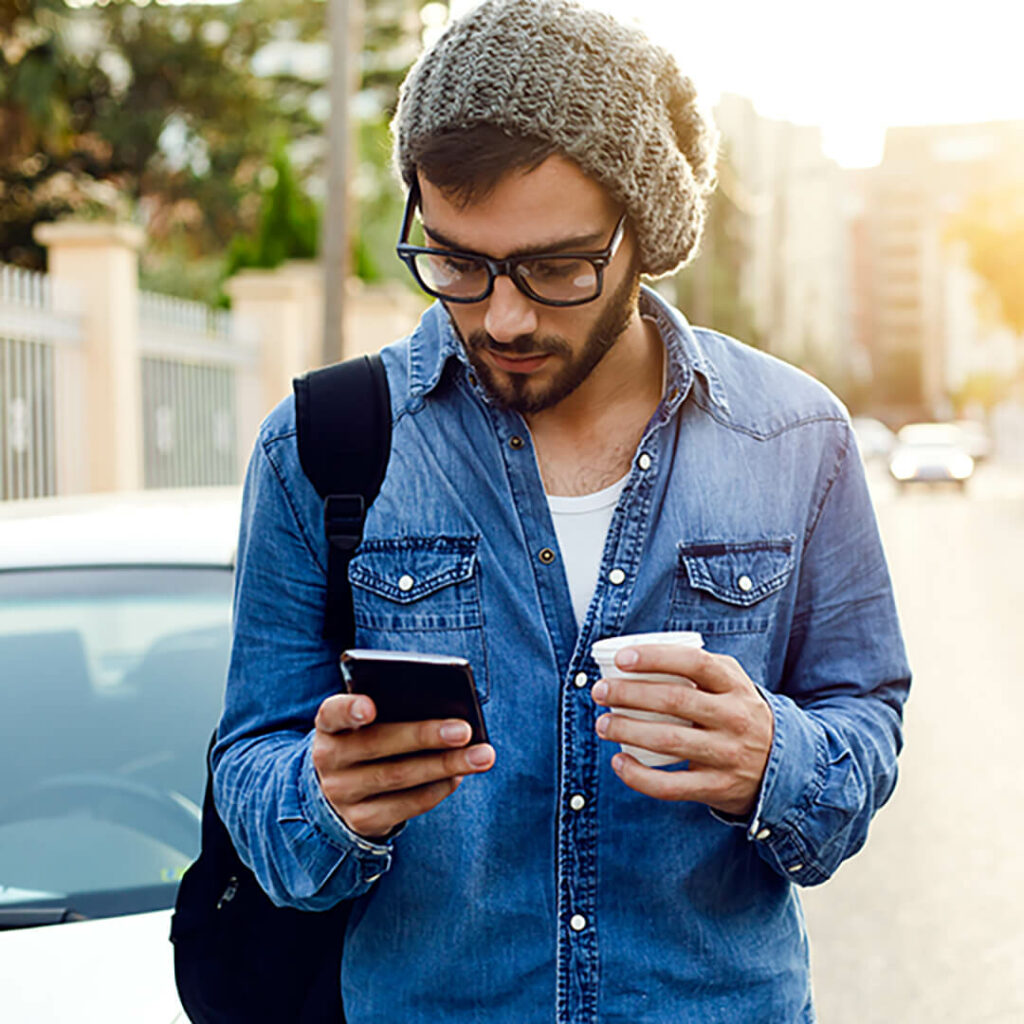 Young man in a jean shirt holding his smartphone in one hand and a to-go cup in the other, while standing on the street next to a car