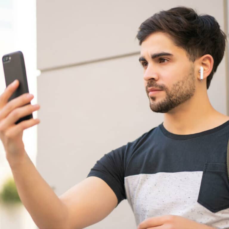 man with dark hair wearing a t-shirt with headphones in holding phone with right hand taking a selfie