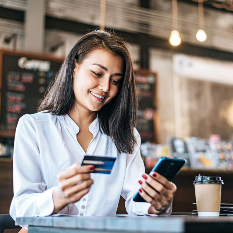 young woman in white blouse sitting in a cafe with her phone in one hand and credit card in the other