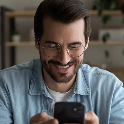 man wearing a denim polo and eyeglasses looking at his phone smiling