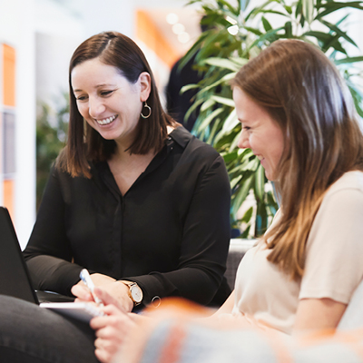 two women smiling sitting on a sofa with a laptop