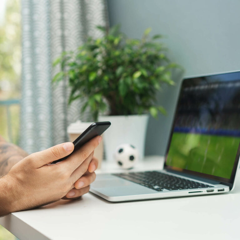hands holding a smartphone in front of a laptop with a soccer game on