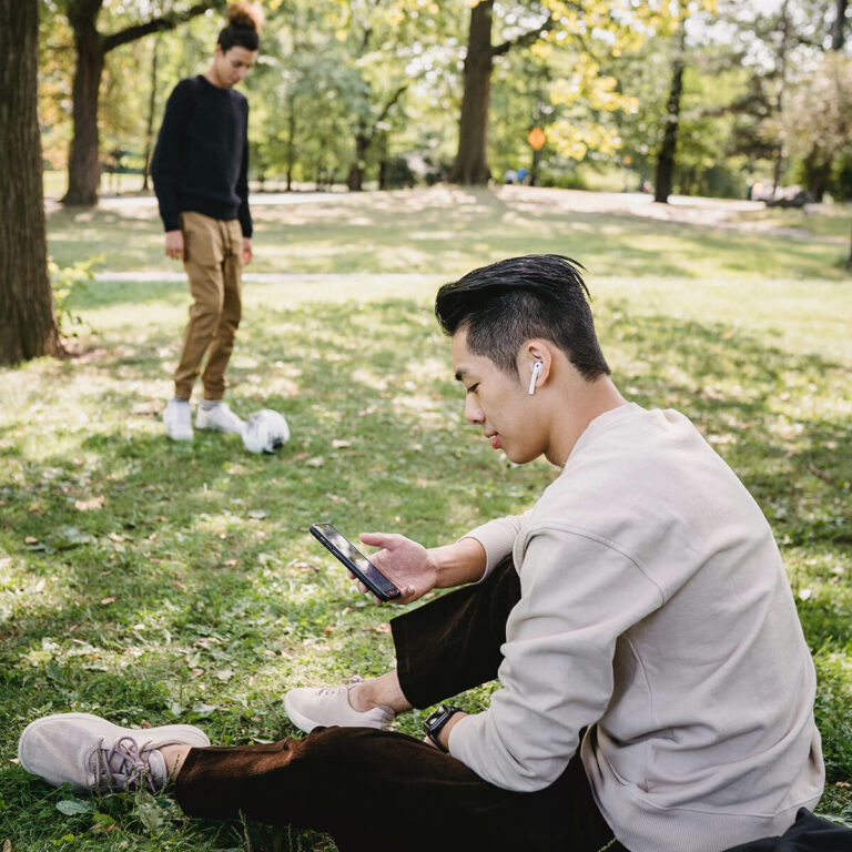 two men in a park with one kicking a soccer ball and the other sitting with headphones in looking at his phone