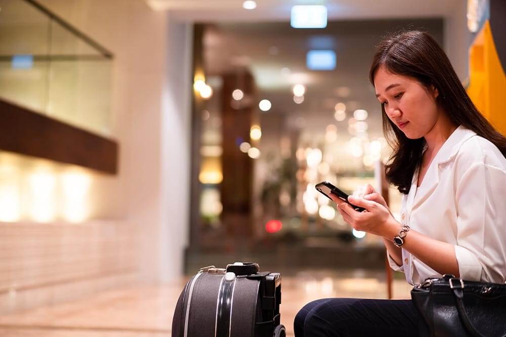 woman sitting in a lobby looking at her phone with her suitcase next to her