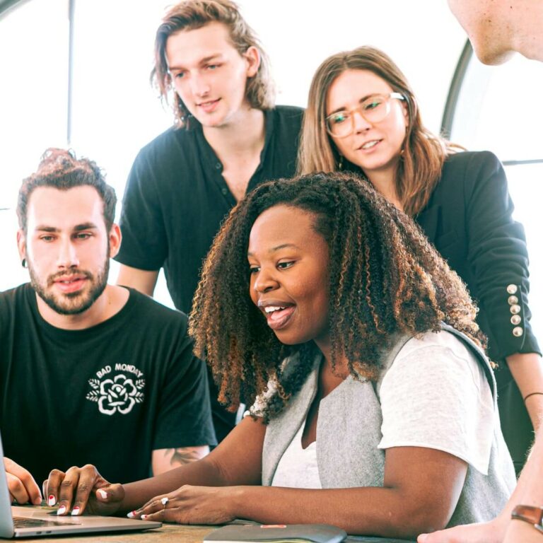 four people standing in front of a laptop and looking at it interested