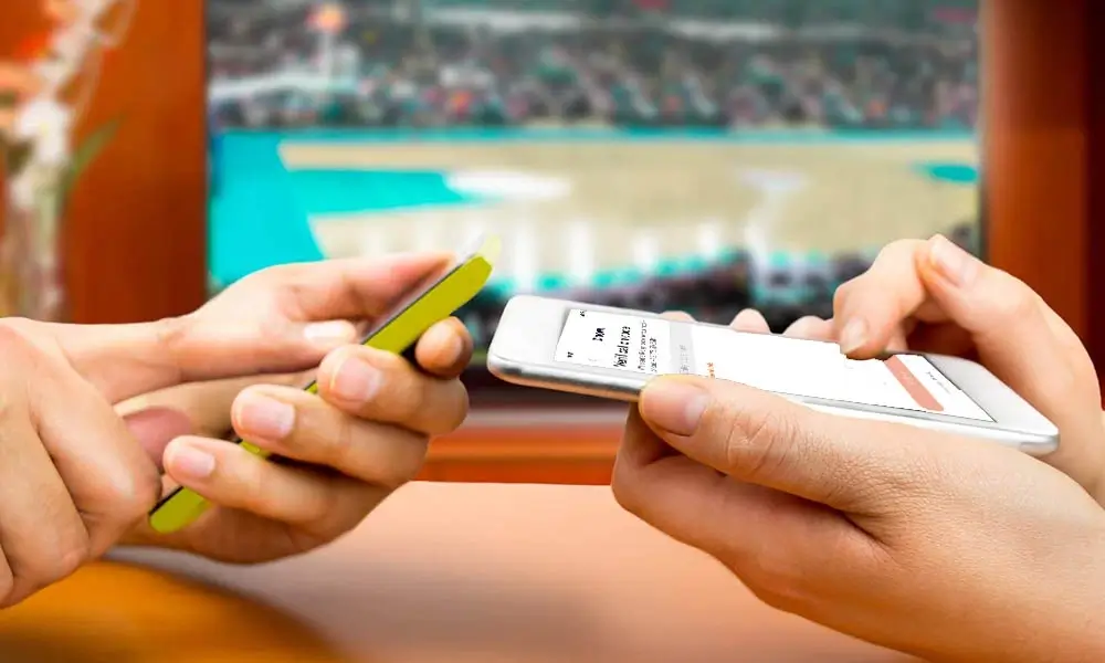 two people holding smartphones in front of a tv with a sports game on